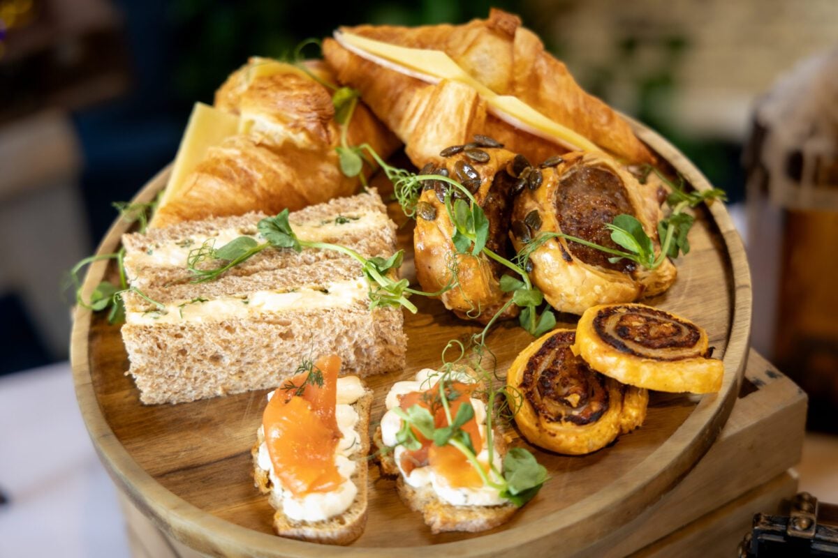 A wooden platter filled with a selection of savoury afternoon tea treats, including sandwiches, croissants with cheese, mini sausage rolls, smoked salmon canapés, and pastry pinwheels, garnished with fresh herbs and microgreens.