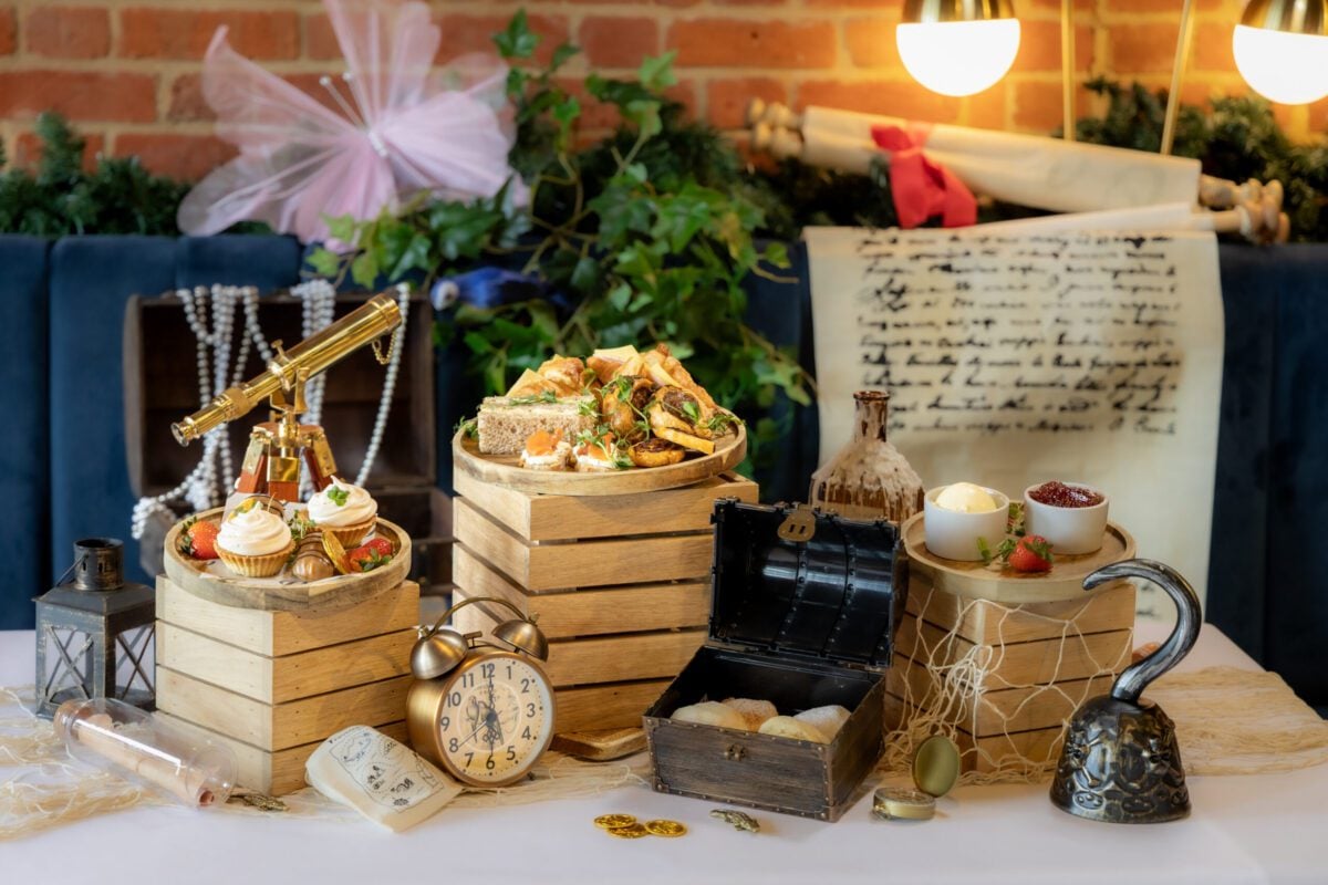 A themed afternoon tea table display featuring sandwiches, cupcakes, scones, and props like a telescope, treasure chest, and Captain Hook’s hook, creating a whimsical pirate and fairy setting.