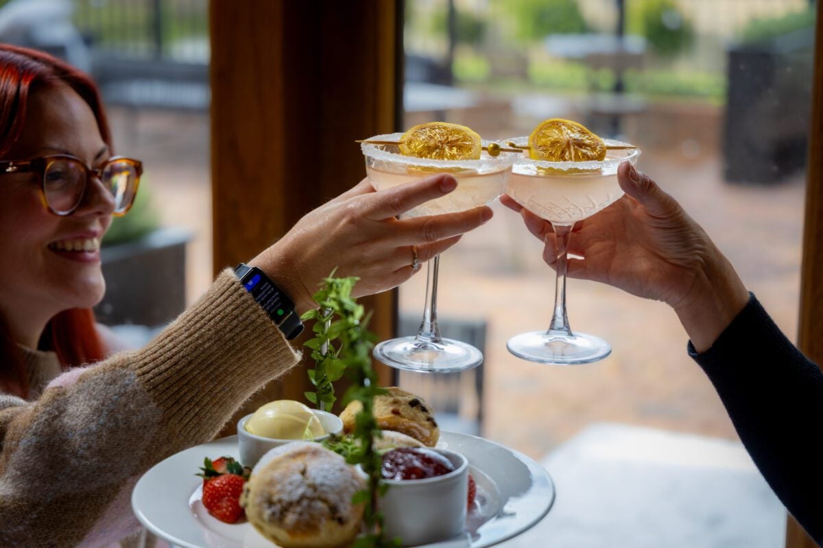 Two adults clinking cocktail glasses garnished with dried lemon slices while enjoying an afternoon tea spread with scones, jam, and strawberries by a window.
