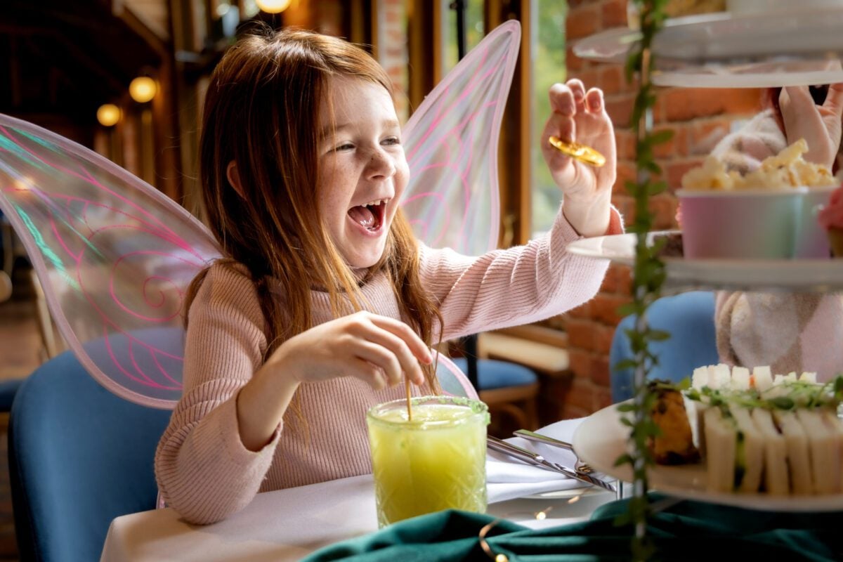 A young girl wearing pink fairy wings laughs with delight while holding a slice of dried lemon and drinking a bright green beverage during an afternoon tea.